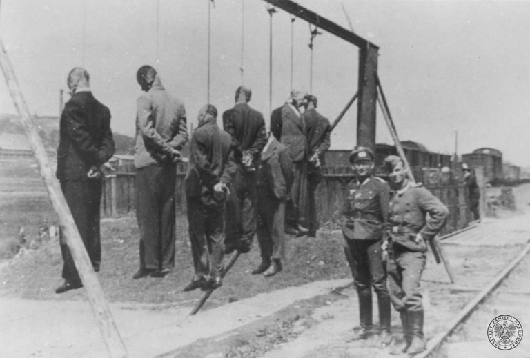 Dwaj Żołnierze Wehrmachtu (oficer i podoficer) pozujący obok szubienicy z ciałami powieszonych Polaków. Zdjęcie wykonał nieznany z nazwiska Niemiec.